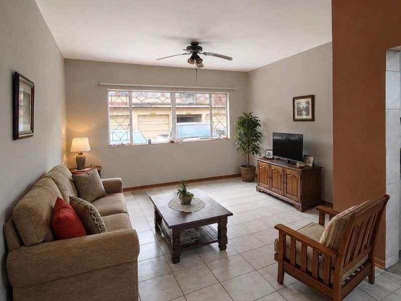 A spacious living room featuring neutral tiled flooring, a ceiling fan, and a large window with security bars, offering a bright and functional living space.
