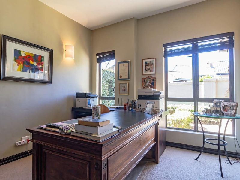 A well-lit home office featuring large windows, neutral wall tones, and carpeted flooring, providing a functional and comfortable workspace.
