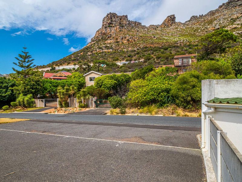 A street-level view of the property entrance, featuring a paved driveway, boundary wall, and established garden set against a mountain backdrop.