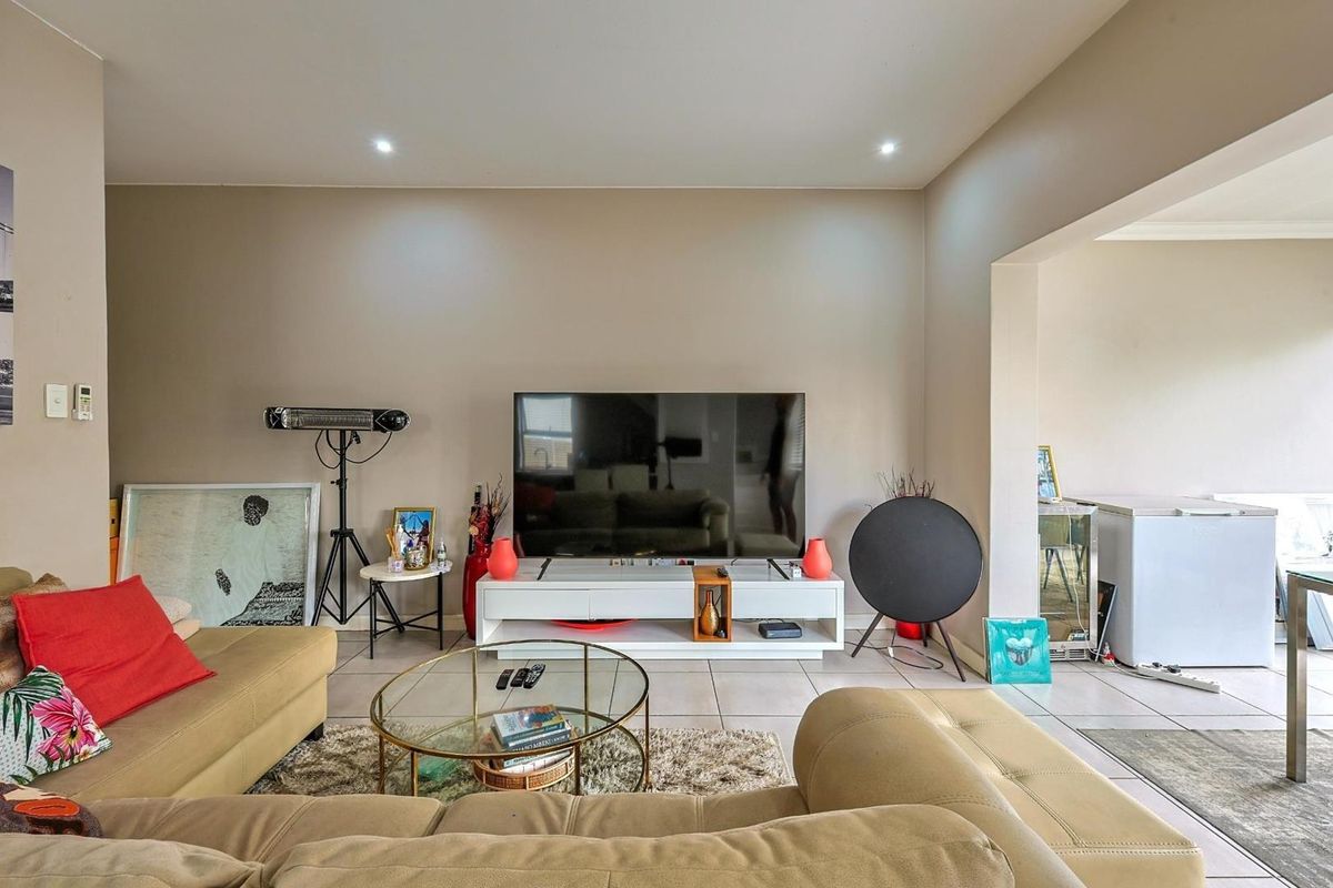 A spacious living room featuring neutral wall tones, recessed downlighting, and large format floor tiles, with an open-plan layout connecting to adjacent areas.