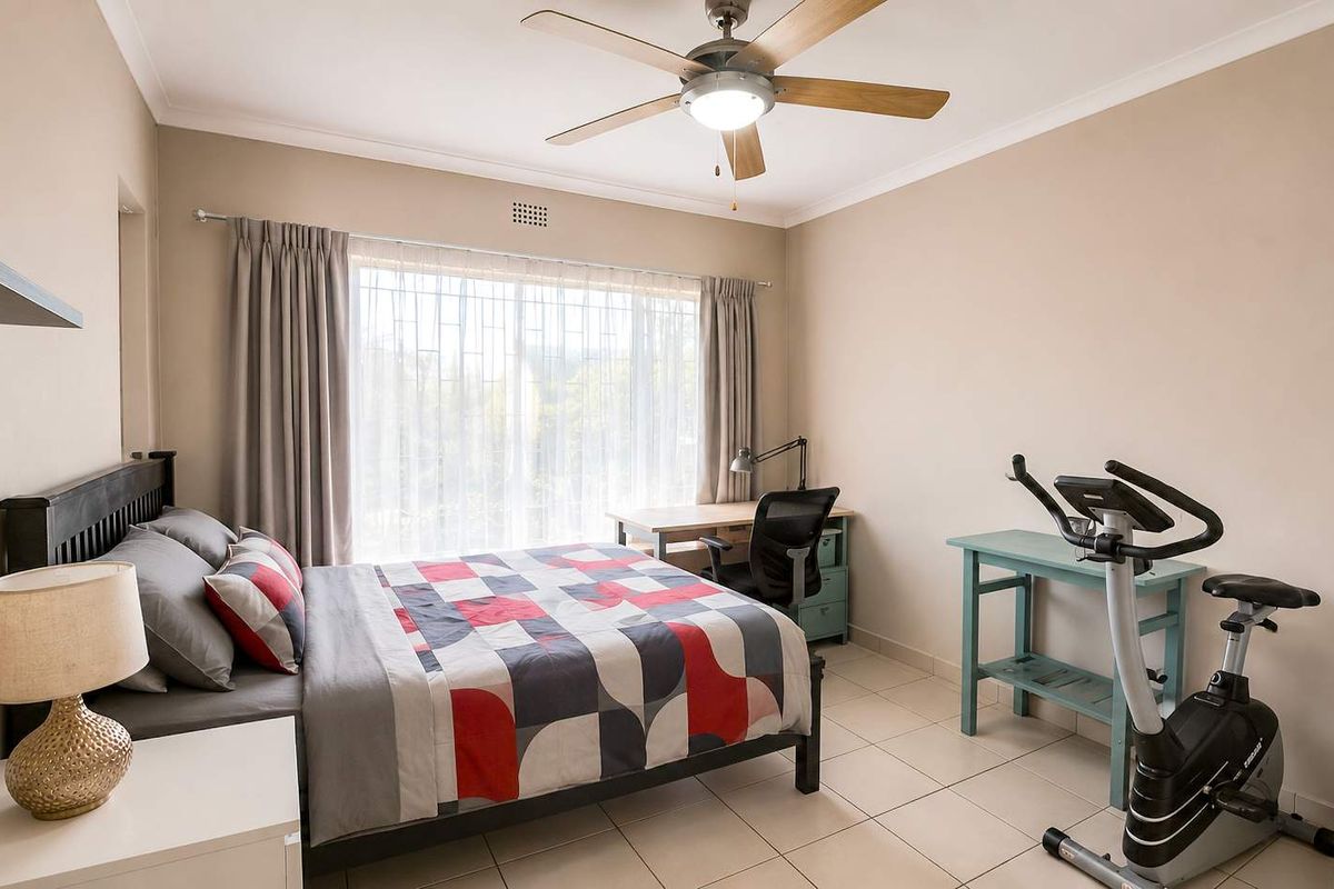 A well-lit bedroom featuring neutral wall tones, tiled flooring, a ceiling fan, and large windows providing natural light.