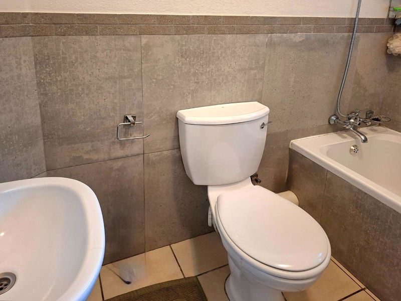 A functional bathroom featuring a standard toilet, bathtub with shower mixer, and basin, finished with neutral wall and floor tiling.