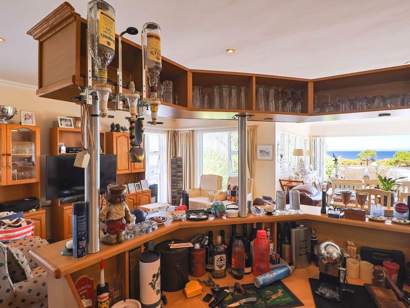 A built-in home bar area featuring wooden cabinetry, overhead glass storage, and recessed lighting, overlooking an open-plan living space with ocean views.