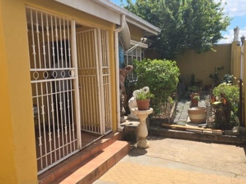 A rear exterior view featuring a paved patio area, security gates, and a side garden walkway enclosed by boundary walls.
