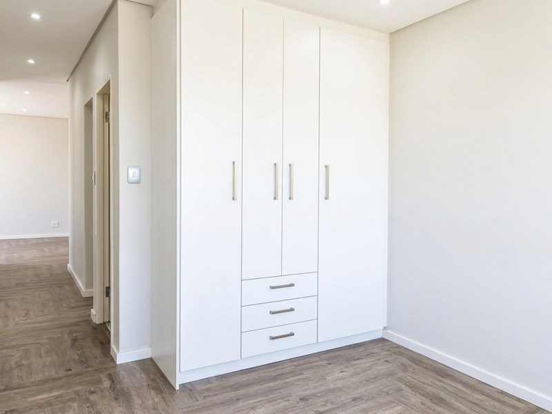A bright, modern bedroom featuring neutral tones, high-quality laminate flooring, and ample built-in cupboard storage with sleek hardware.