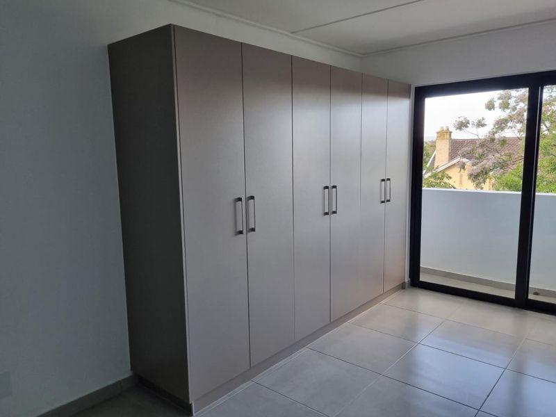 A bright bedroom featuring modern built-in cupboards, neutral tiled flooring, and a sliding glass door providing access to a private balcony.