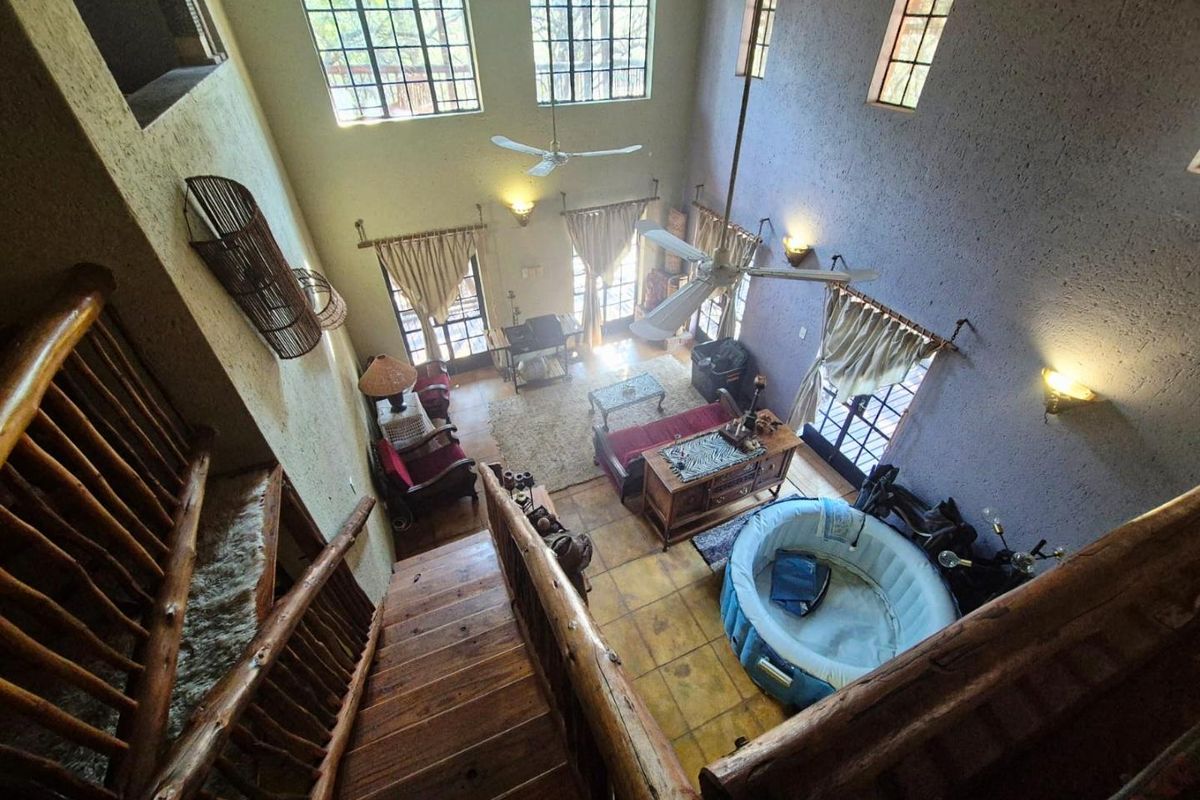 Spacious double-volume living area featuring high ceilings, large windows, rustic timber staircase, and tiled flooring with direct access to the outdoor area.