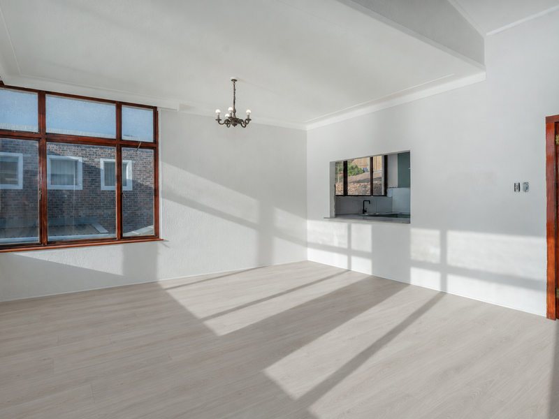 Bright and spacious living area featuring light-toned laminate flooring, large timber-framed windows, and a convenient serving hatch connecting to the kitchen area.