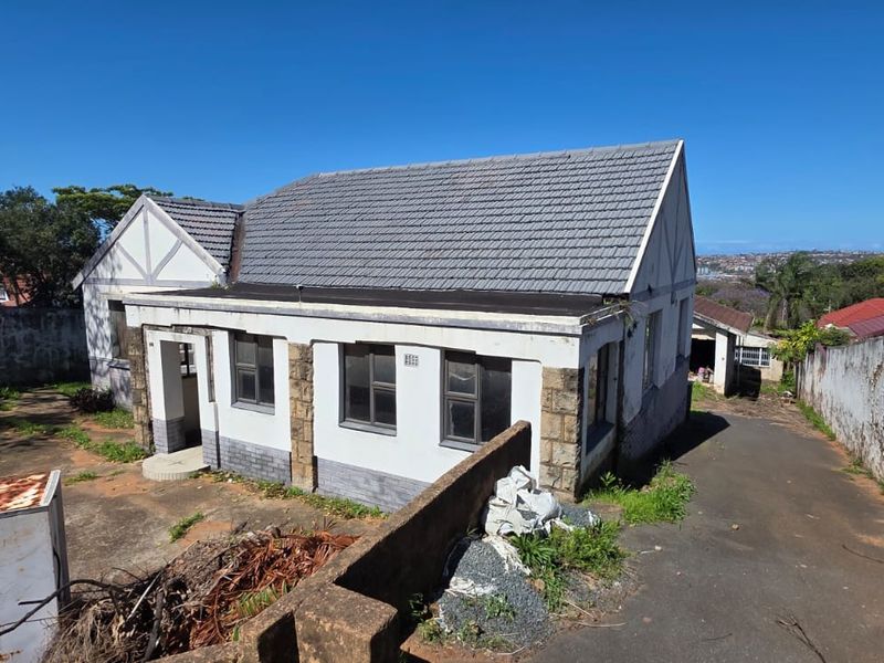 Rear view of a detached house featuring a tiled roof, stone accents, and a paved driveway, requiring significant exterior maintenance and landscaping.