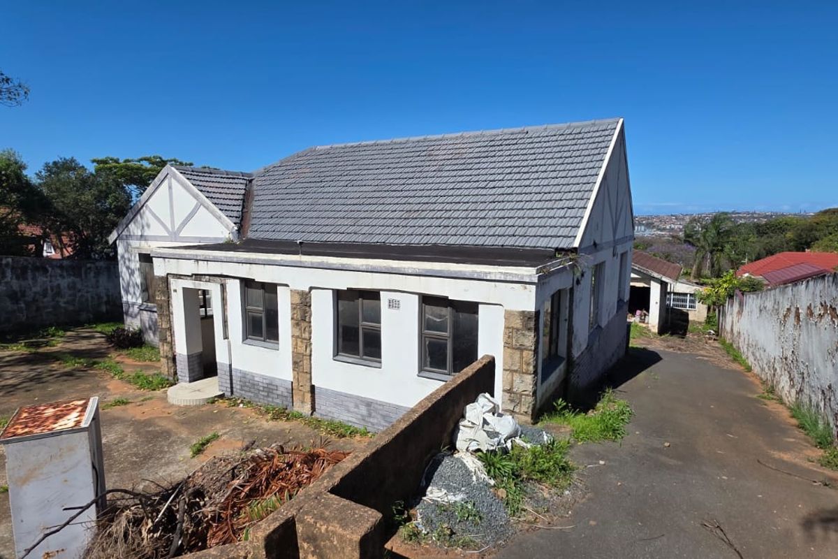 Rear view of a detached house featuring a tiled roof, stone accents, and a paved driveway, requiring significant exterior maintenance and landscaping.