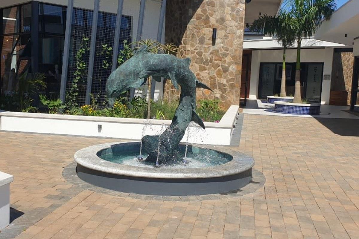 A well-maintained paved courtyard featuring a central dolphin water fountain, stone-clad building exterior, and landscaped garden beds.