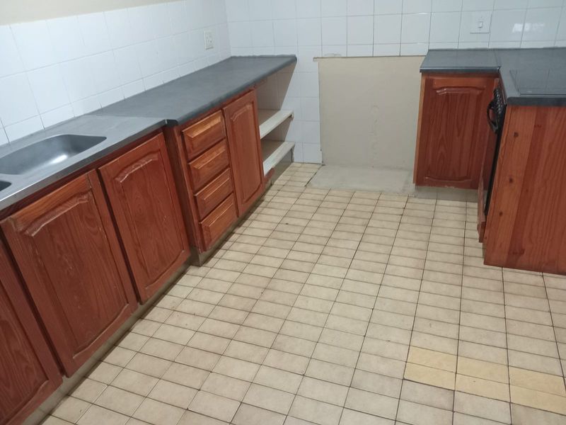 A dated kitchen featuring wooden cabinetry, laminate countertops, and tiled flooring, currently vacant and requiring cosmetic updates to modernize the space.