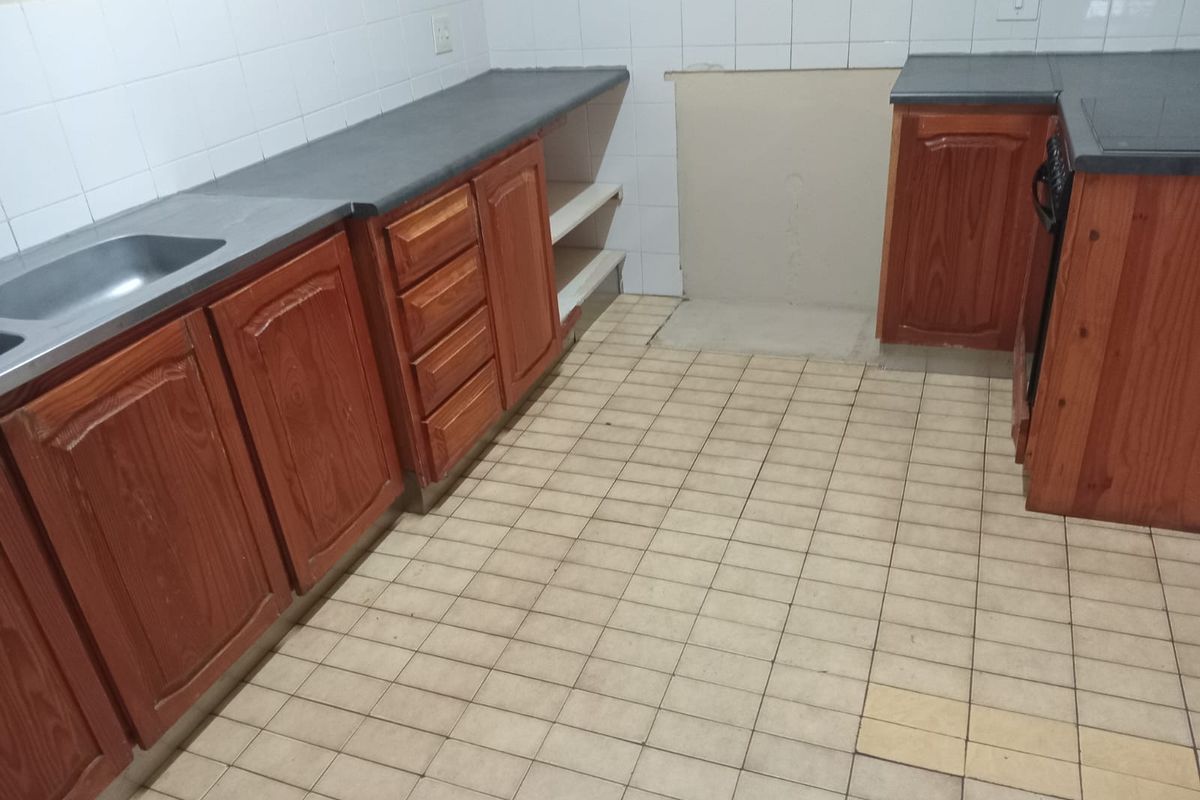 A dated kitchen featuring wooden cabinetry, laminate countertops, and tiled flooring, currently vacant and requiring cosmetic updates to modernize the space.