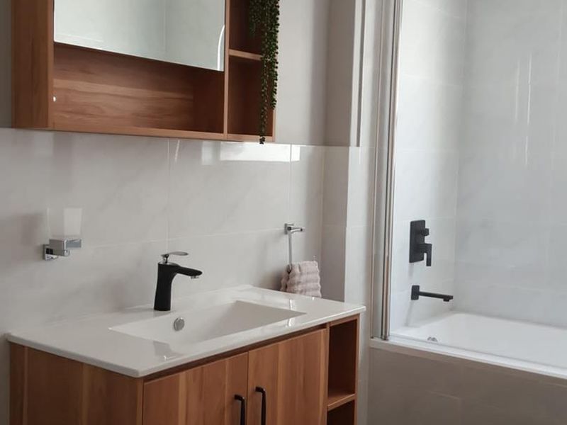 Modern bathroom featuring a wood-finish vanity with integrated basin, black fixtures, and a bathtub with a glass shower screen and rainfall shower head.