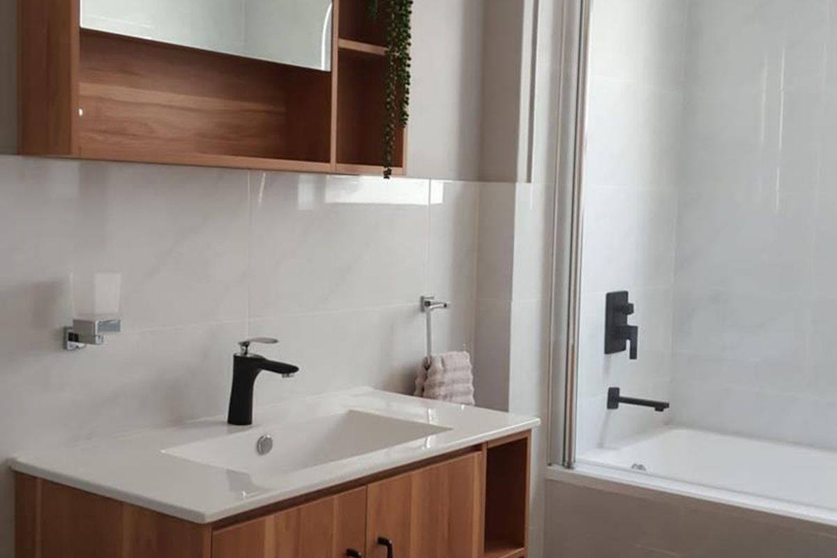 Modern bathroom featuring a wood-finish vanity with integrated basin, black fixtures, and a bathtub with a glass shower screen and rainfall shower head.