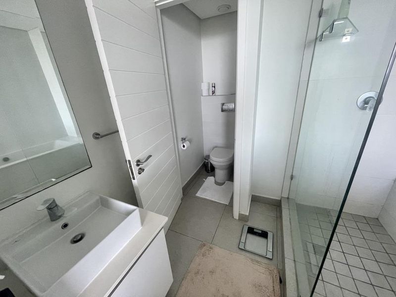 A modern bathroom featuring a vanity with basin, a glass-enclosed shower, and a separate toilet area with neutral tiling.