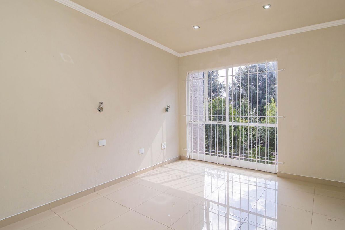 A bright, empty bedroom featuring neutral tiled flooring, recessed ceiling lighting, and a large window secured with white metal burglar bars.