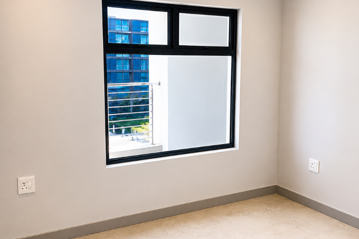 A bright, empty bedroom featuring neutral tiled flooring, recessed downlighting, and a modern black-framed window with a view of the surrounding apartment complex.