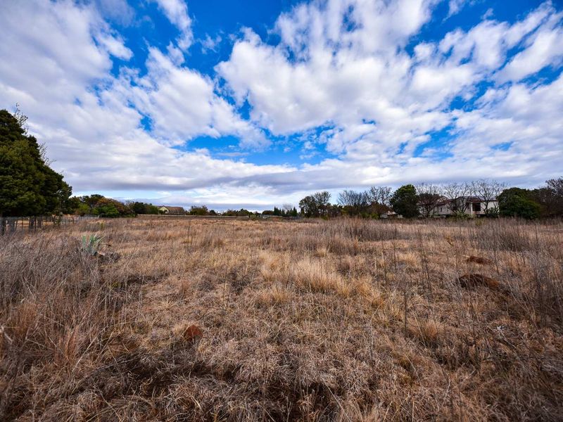 A wide-angle view of a vacant residential plot featuring dry grass, mature trees, and perimeter wire fencing under a bright blue sky.