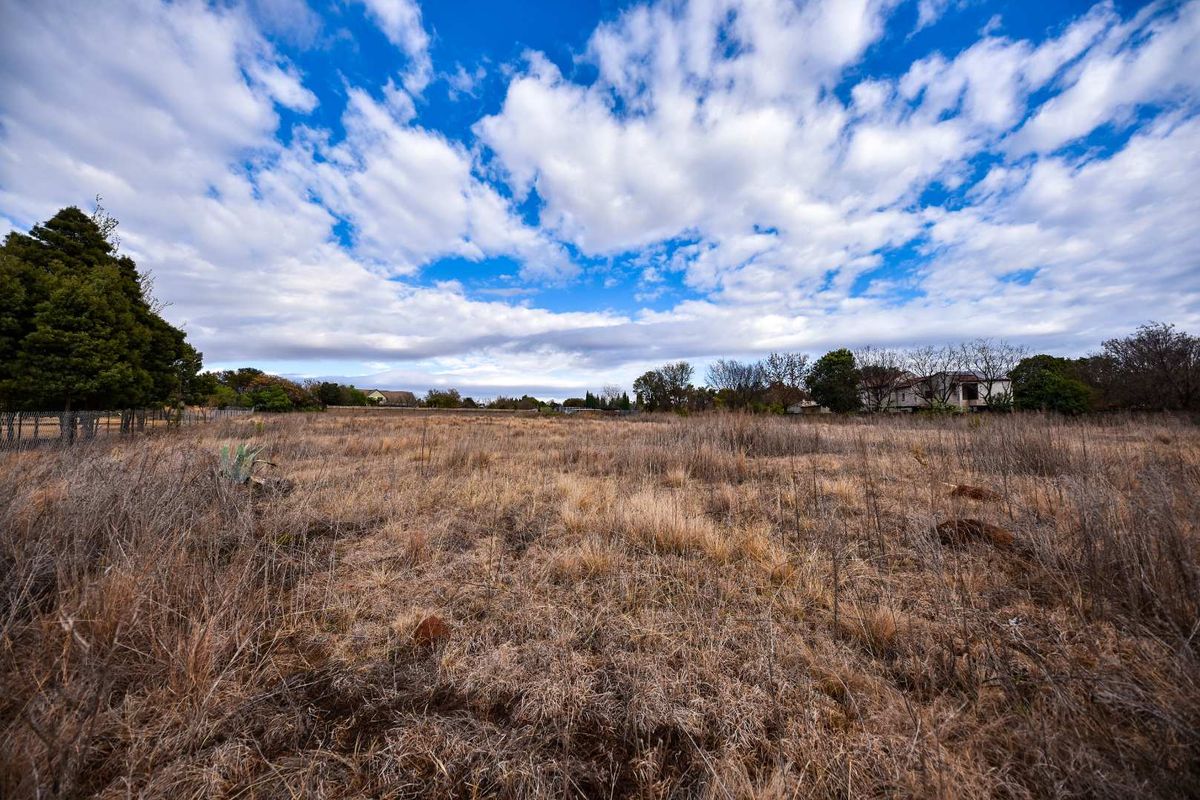 A wide-angle view of a vacant residential plot featuring dry grass, mature trees, and perimeter wire fencing under a bright blue sky.