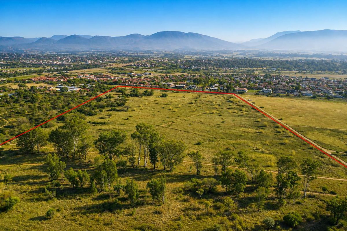 An aerial view of a large, undeveloped farm plot with a red boundary line indicating the property perimeter, set against a mountain landscape.