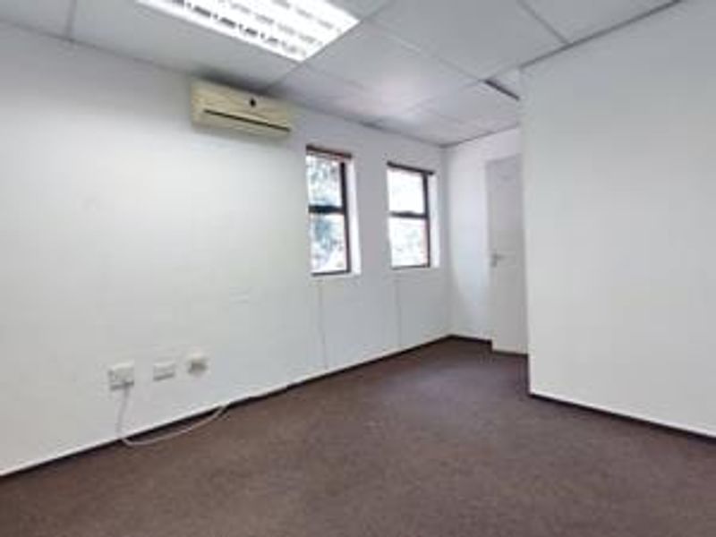 An empty office space featuring a drop ceiling, fluorescent lighting, wall-mounted air conditioning, and carpeted flooring.