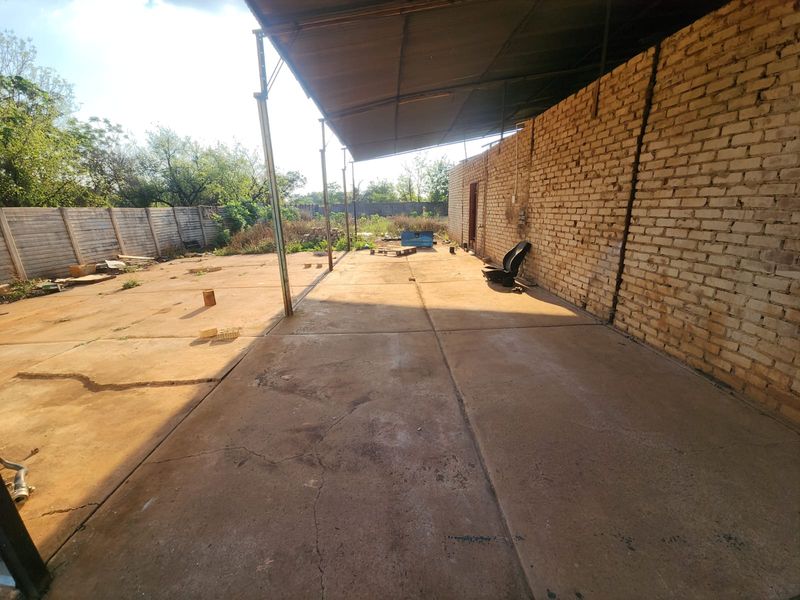 An expansive outdoor covered patio area featuring a concrete slab floor, exposed brick wall, and metal roof structure, offering significant potential for renovation.