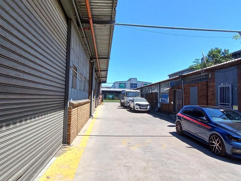 Industrial exterior view showing a concrete driveway between brick warehouse buildings with large roller shutter doors and secure access points.