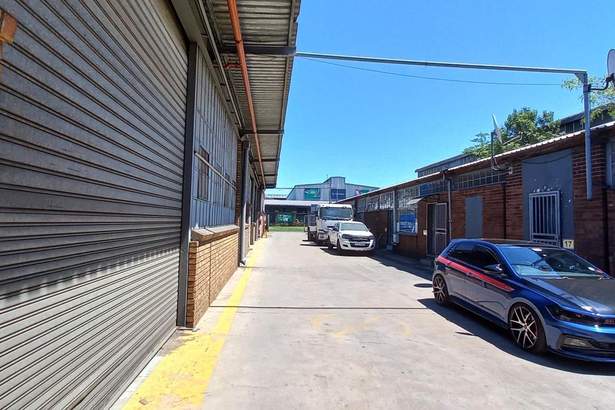 Industrial exterior view showing a concrete driveway between brick warehouse buildings with large roller shutter doors and secure access points.
