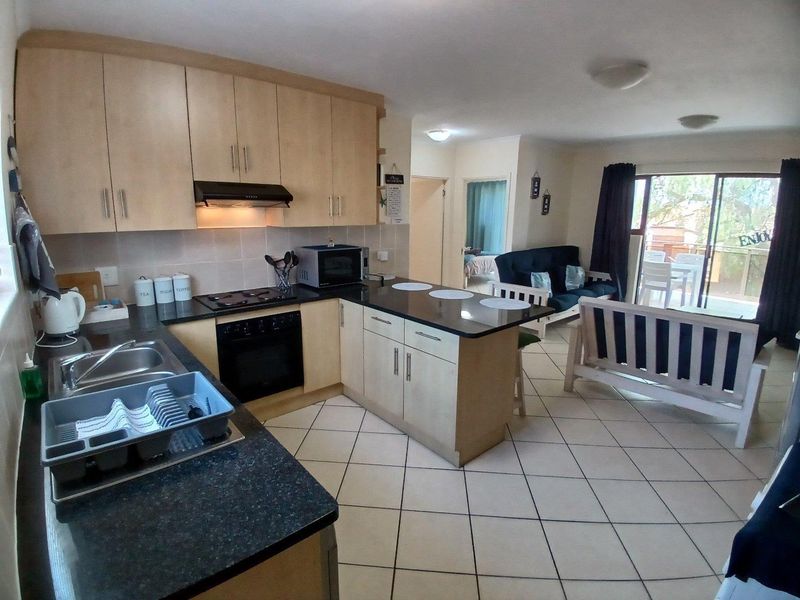 Open-plan kitchen and living area featuring light wood cabinetry, dark countertops, tiled flooring, and direct access to a balcony through sliding glass doors.