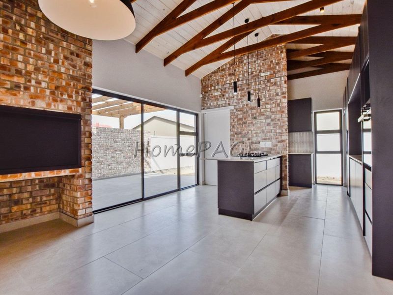 Spacious open-plan living and kitchen area featuring exposed brick walls, timber ceiling beams, a modern kitchen island with gas hob, and large sliding glass doors.