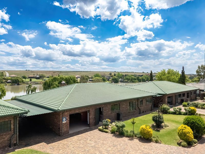 Aerial view of a brick house with a tiled roof, paved driveway, and landscaped garden, overlooking a scenic lake and surrounding landscape.