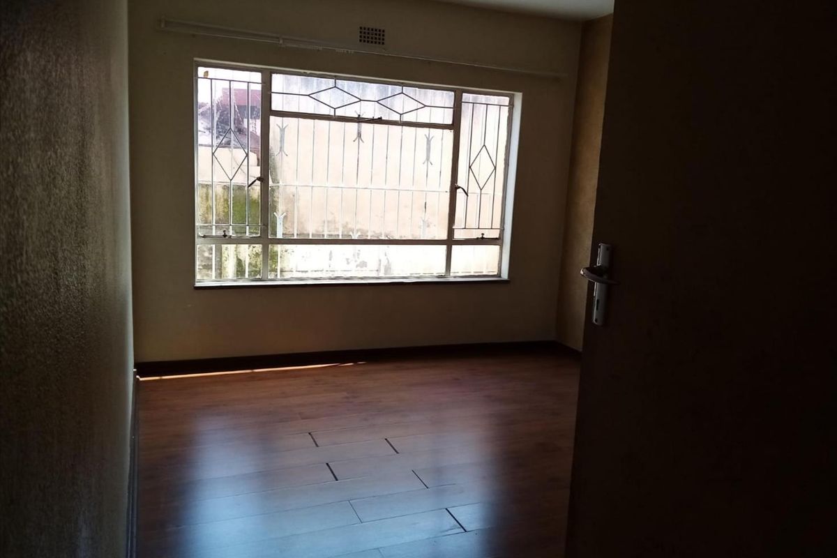 An empty bedroom featuring laminate flooring, a large window with security bars, and a ventilation grille, requiring minor repairs to the flooring.