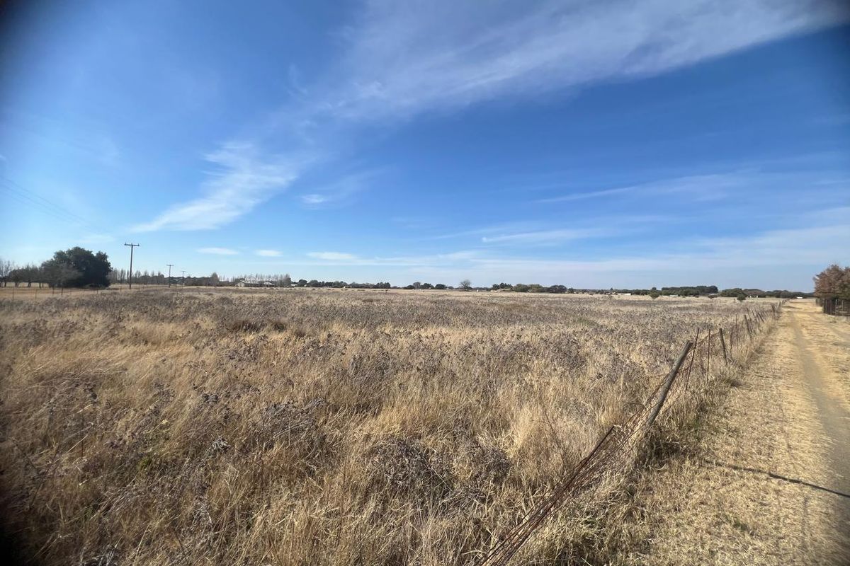 A wide-angle view of a vacant residential plot featuring dry grass, wire fencing, and an adjacent dirt access track under a clear blue sky.