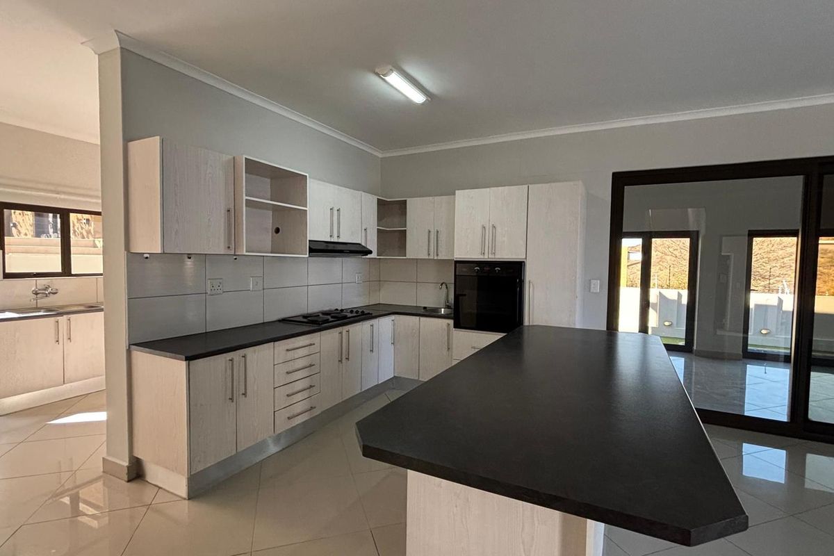 Modern kitchen featuring light wood-finish cabinetry, dark countertops, a central island, built-in oven and hob, and tiled flooring with access to an outdoor area.