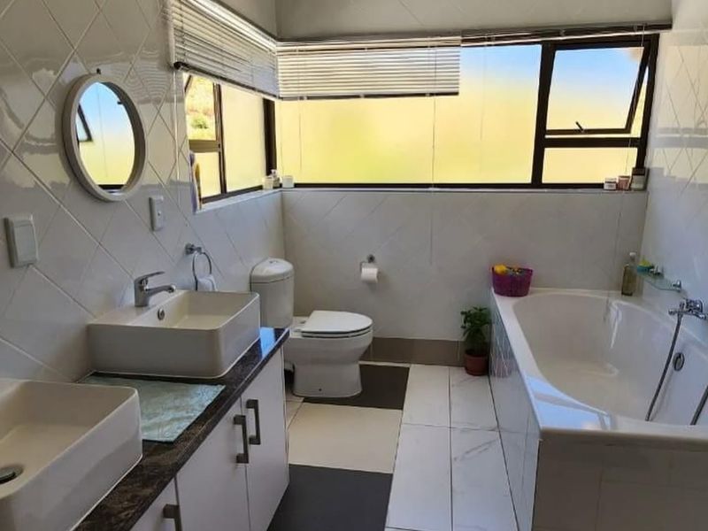 A well-lit bathroom featuring a double vanity, built-in bathtub, toilet, and modern tiled finishes with large corner windows providing natural light.
