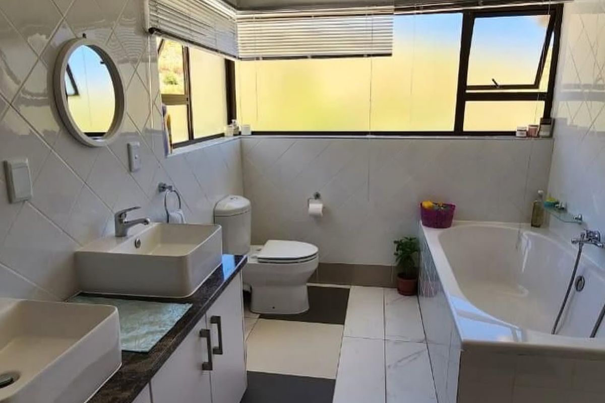 A well-lit bathroom featuring a double vanity, built-in bathtub, toilet, and modern tiled finishes with large corner windows providing natural light.