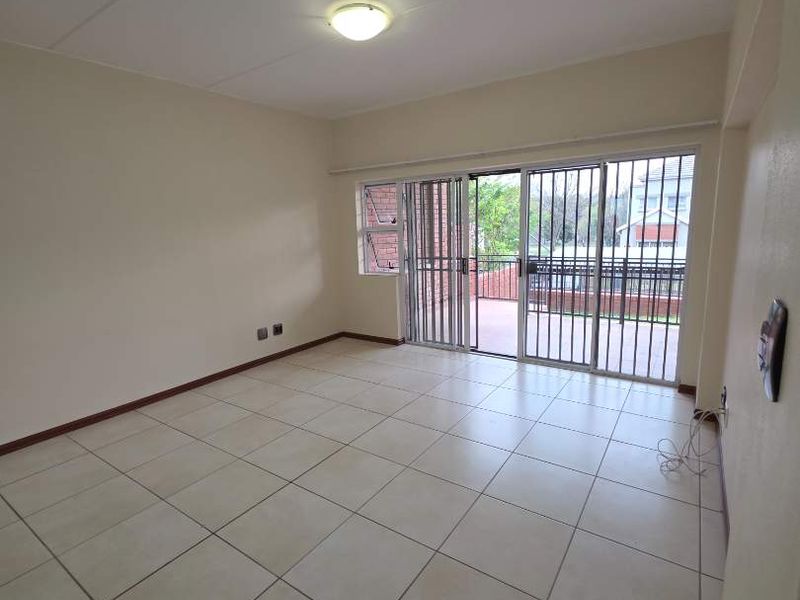 A bright, empty living room featuring neutral tiled flooring and a sliding glass door with security gate leading to an outdoor balcony area.