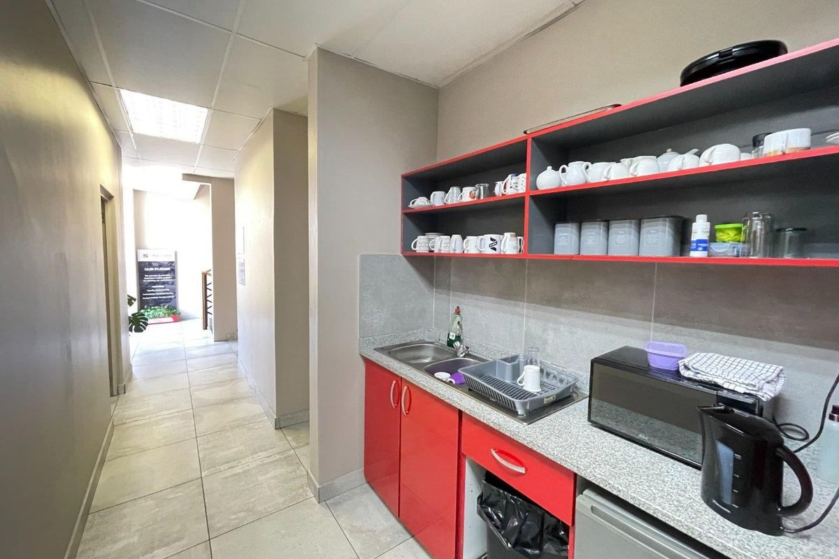 A compact kitchenette area featuring red cabinetry, stone-look countertops, a stainless steel sink, and tiled backsplash, situated within a commercial office hallway.