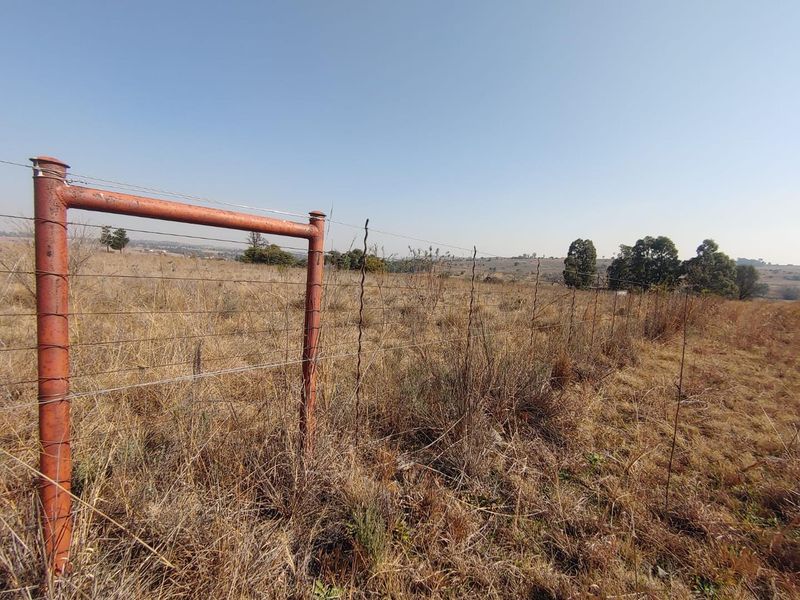 A view of an open agricultural land plot featuring wire fencing and a metal post, with dry grass and distant trees.