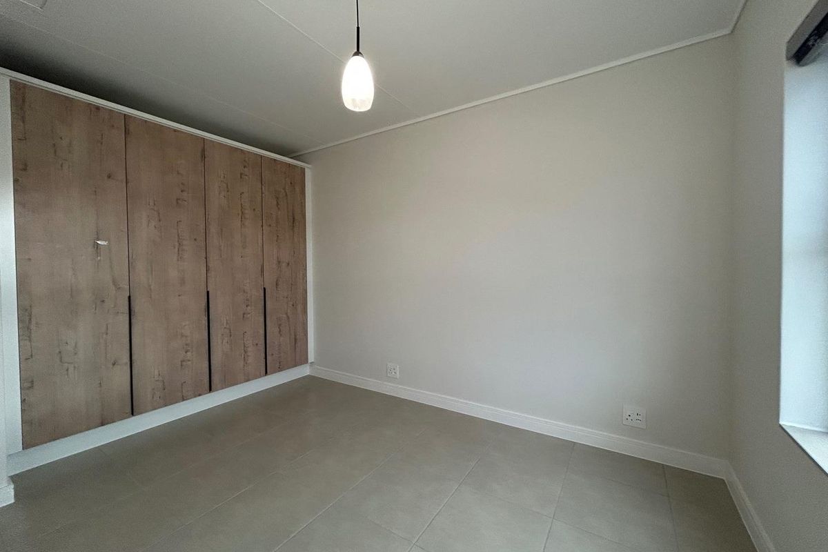 A bright, empty bedroom featuring neutral floor tiles, clean white walls, and modern built-in wooden cabinetry.