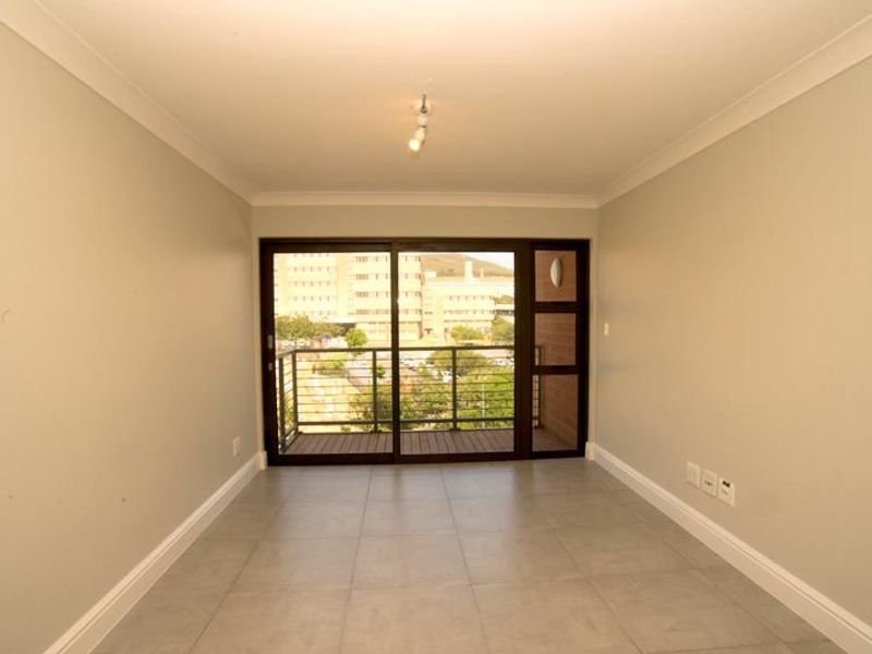 An empty, well-lit living area featuring neutral tiled flooring, cove cornicing, and a large sliding glass door providing access to a private balcony.