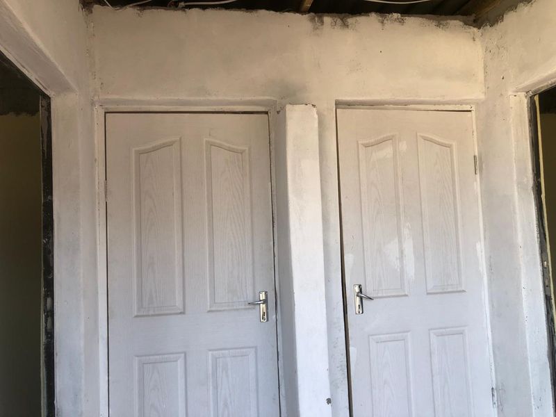 A hallway area featuring two white moulded panel doors with unfinished wall surfaces and exposed ceiling structure, requiring completion and cosmetic finishing.