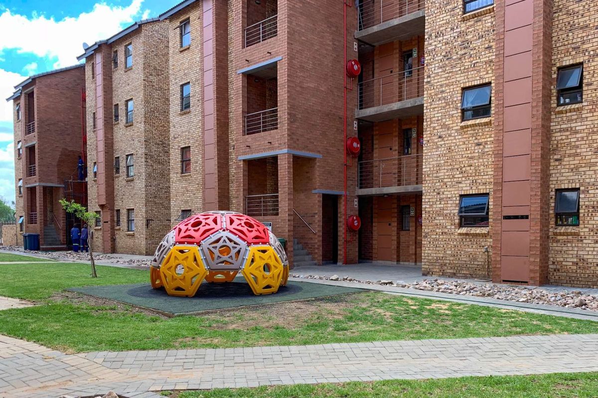 Exterior view of a multi-story apartment complex featuring face brick construction, private balconies, and a communal garden area with a playground structure.