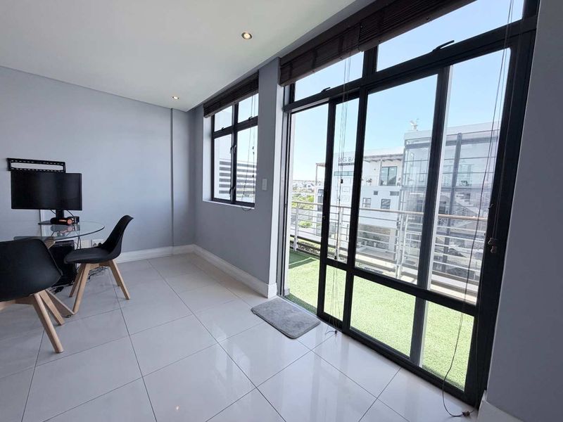 A bright office space featuring large aluminium-framed windows, sliding glass doors leading to a balcony, and modern tiled flooring.