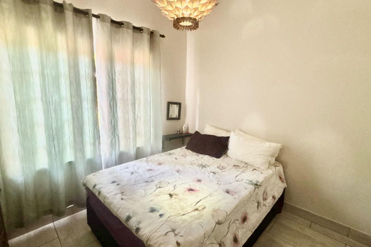 A bedroom featuring tiled flooring and a central ceiling light fixture, with natural light entering through a large window.