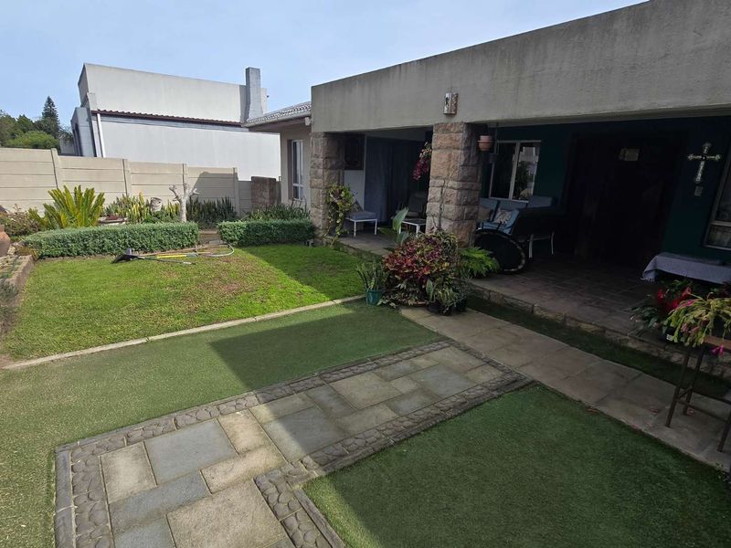 Rear garden area featuring a covered patio with stone-clad pillars, paved pathways, and a mix of natural and artificial lawn surfaces.
