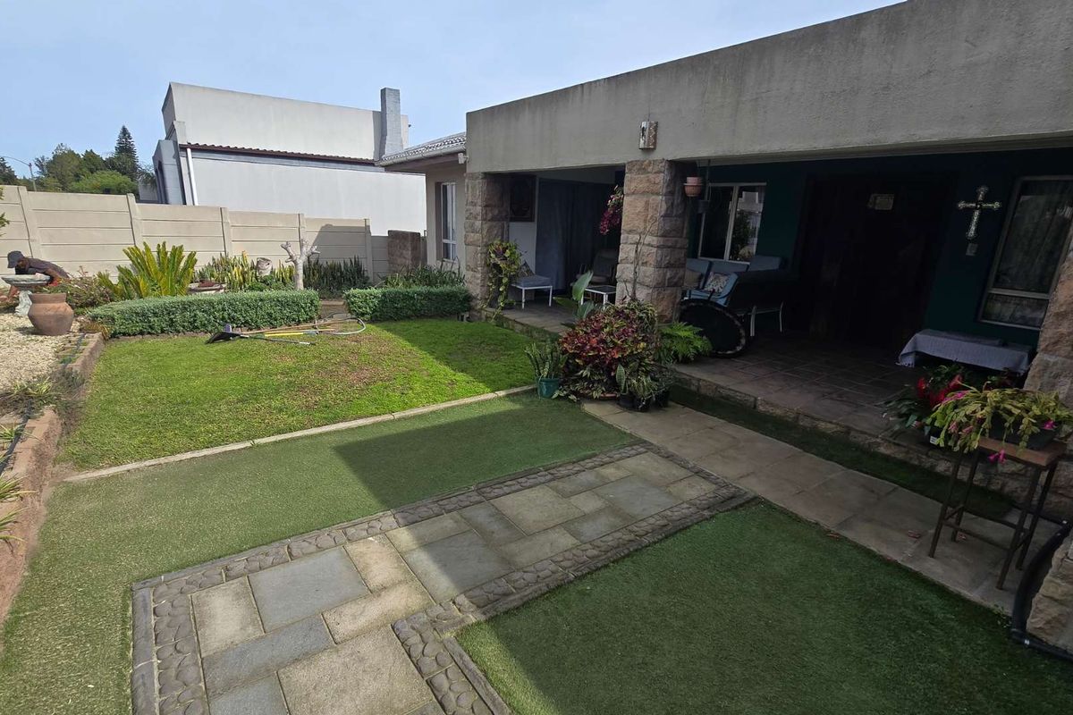 Rear garden area featuring a covered patio with stone-clad pillars, paved pathways, and a mix of natural and artificial lawn surfaces.