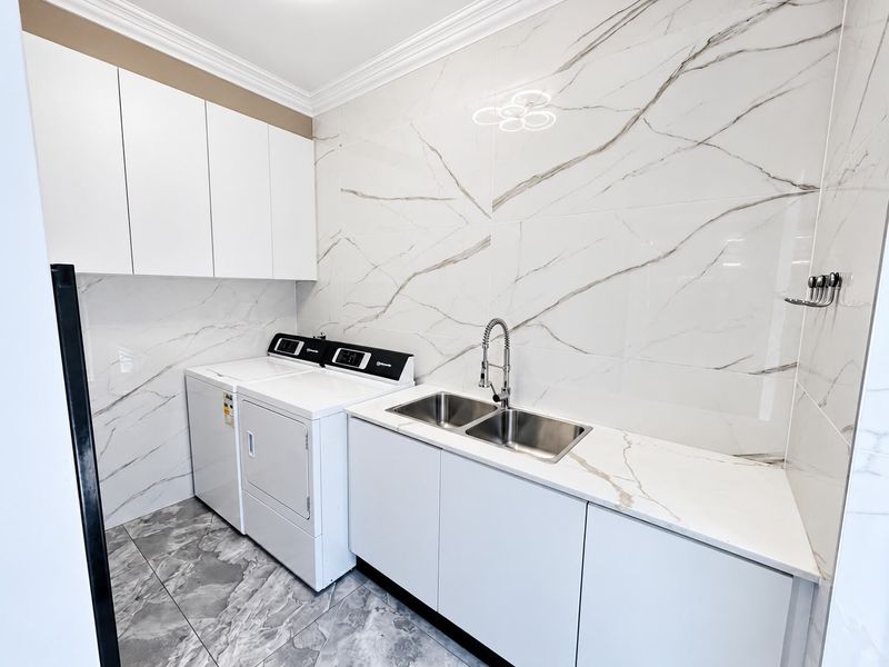 Modern laundry room featuring marble-look wall and floor tiles, sleek white cabinetry, stone-look countertops, and a double stainless steel sink with a gooseneck mixer.