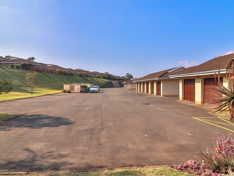 A wide view of the apartment complex exterior featuring a large asphalt driveway and a row of individual garage units under a tiled roof.